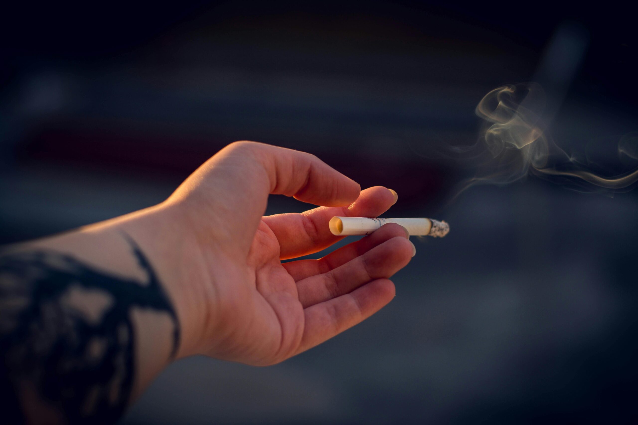 A tattooed hand holding a smoking cigarette, captured in a close-up with shallow focus.